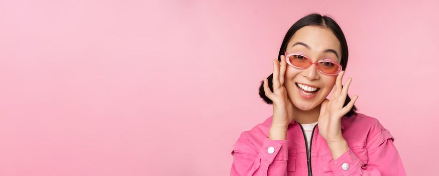 Cute modern japanese girl in sunglasses, smiling and looking happy, posing against pink background in stylish clothing.