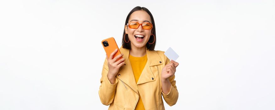 Online shopping and delivery concept. Happy korean girl in stylish clothes, holding credit card and smartphone, laughing and smiling, standing over white background.