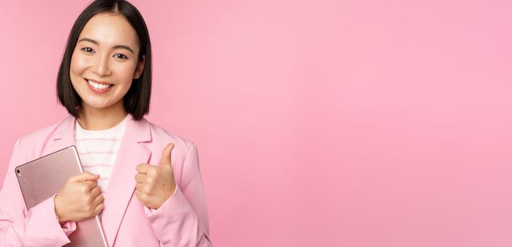 Portrait of corporate woman, girl in office in business suit, holding digital tablet, showing thumbs up, recommending company, standing over pink background.