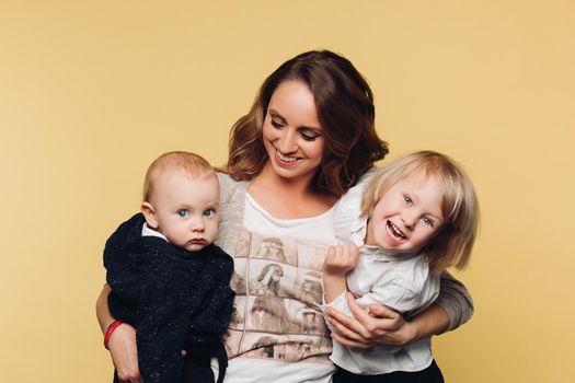 Happy young mother posing at camera together with her kids. Beautiful parent in casual clothes holding little baby and giving hand to other one. Brunette woman smiling and standing with cute sons.