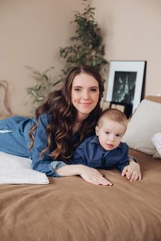 Portrait of gorgeous brunette young woman in casual denim dress embracing her lovely son in denim shirt on the bed. Family and motherhood concept.