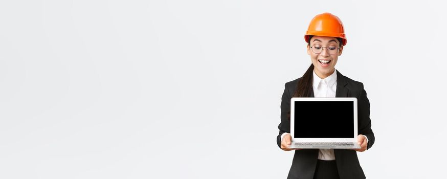 Impressed happy asian female chief engineer showing great results of enterprise to investors, standing in safety helmet and business suit, holding laptop and look pleased at computer screen.