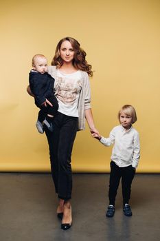 Happy young mother posing at camera together with her kids. Beautiful parent in casual clothes holding little baby and giving hand to other one. Brunette woman smiling and standing with cute sons.