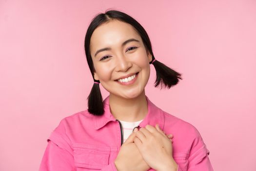 Cute korean girl with silly hairbuns, looking happy and grateful, thank you pose, holding hands on heart flattered, posing against pink background.