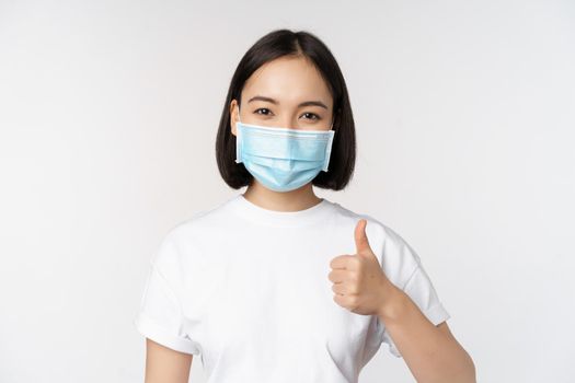 Smiling asian girl in medical mask showing thumbs up, approve something good, praise and compliment company, standing over white background.