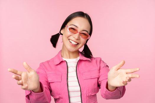 Happy stylish korean girl in sunglasses, reaching hands, receiving, smiling friendly and welcome you, standing over pink background.