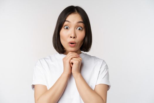 Close up portrait of asian brunette woman looking amazed, say wow, watching smth impressive, standing over white background.