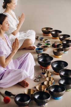 Two women are sitting with Tibetan bowls in the lotus position before a yoga class in the gym.