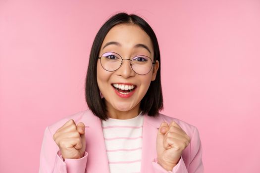 Close up portrait of asian businesswoman cheering, rooting for, looking with hope and excitement at camera, smiling and laughing, standing over pink background.