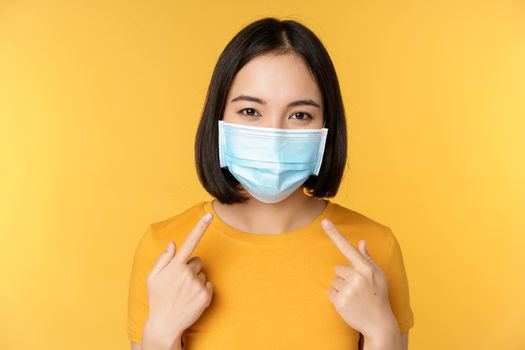 Portrait of smiling asian woman in medical face mask, pointing at her personal protective equipment from covid-19 during pandemic, standing against yellow background.