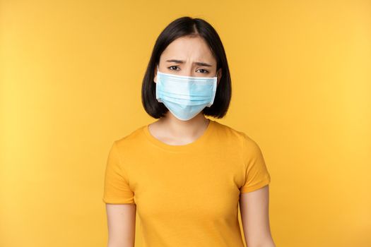 Image of sad and disappointed asian woman in medical mask, frowning and looking displeased, standing against yellow background.