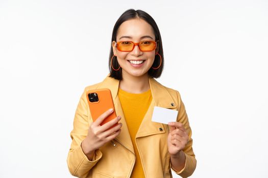 Online shopping. Stylish asian female model in sunglasses, holding credit card and mobile phone, smiling happy, standing over white background. Copy space