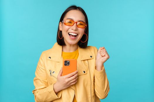 Enthusiastic smiling asian girl in sunglasses, holding mobile phone and dancing, laughing with smartphone, standing over blue background.