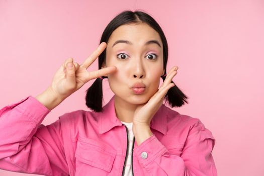 Close up portrait of stylish asian girl shows peace, v-sign and kissing, posing against pink background.