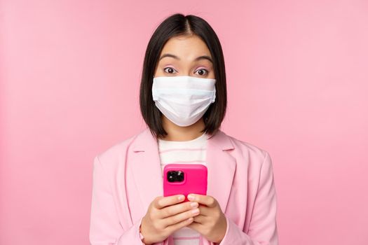 Business people and covid-19 concept. Young asian businesswoman in suit and medical face mask, using smartphone app, standing over pink background.