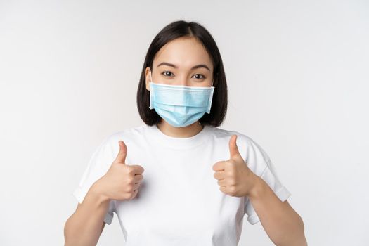 Covid-19, health and people concept. Young happy korean woman in medical mask, showing thumbs up, standing in tshirt over white background.