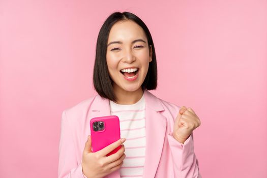 Enthusiastic asian businesswoman saying yes, winning on mobile phone, using smartphone and triumphing, celebrating success, standing over pink background.