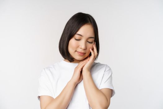 Portrait of tender and feminine young asian woman, looking down, gently touching face, concept of skin care and cosmetology, white background.