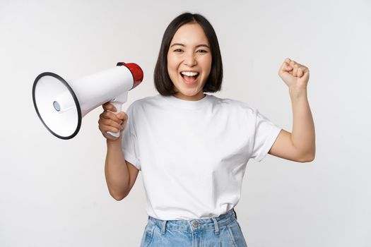 Happy asian woman shouting at megaphone, making announcement, advertising something, standing over white background.