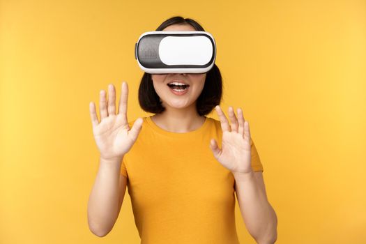 Young asian woman playing in VR, using virtual reality glasses, wearing yellow t-shirt against studio background. Copy space