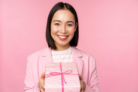 Smiling asian businesswoman in suit, giving you gift in wrapped box, standing over pink background. Copy space