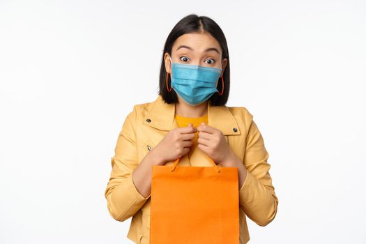 Covid-19 and shop concept. Young asian stylish woman, wearing medical face mask, holding shopping bag, going in malls during pandemic, white background.