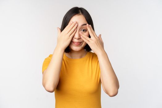 Cute korean female model, cover eyes with hands, peeking through fingers curious, watching smth, standing in yellow tshirt over white background.