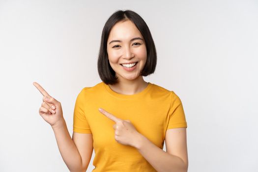 Portrait of smiling asian brunette girl in yellow tshirt, pointing fingers left, showing copy space, promo deal, demonstrating banner, standing over white background.