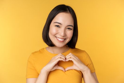 Close up portrait of smiling korean woman, showing romantic heart sign and looking happy, standing over yellow background. Copy space