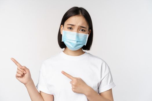 Sad asian woman in medical mask, pointing fingers left, frowning and looking upset, complaining, demonstrating banner, standing over white background.