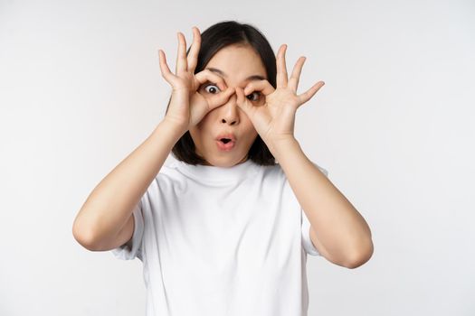 Funny young asian woman, korean girl making eyes glasses gesture, looking happy at camera, standing over white background.