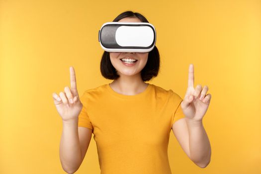 Young asian woman playing in VR, using virtual reality glasses, wearing yellow t-shirt against studio background. Copy space