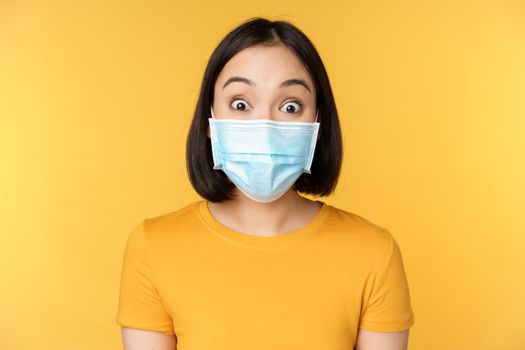 Covid-19 and medical concept. Close up portrait of asian woman in face mask, looking surprised and amazed at news, standing over yellow background.