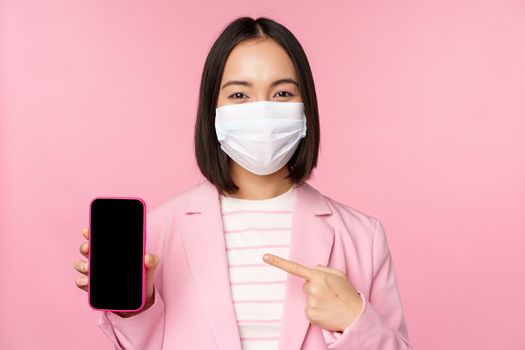 Smiling korean businesswoman in suit, wearing medical face mask from covid, pointing finger at mobile phone screen, standing over pink background.