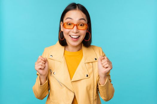 Image of enthusiastic young asian woman celebrating, triumphing, looking surprised and happy, clapping hands satisfied, standing over blue background.