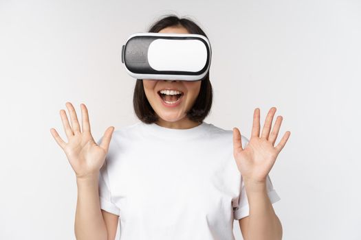 Happy asian woman using VR headset, waving raised hands and laughing, using virtual reality glasses, standing over white background.