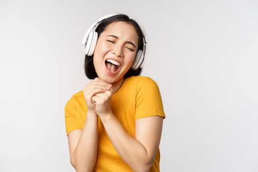 Happy asian girl dancing, listening music on headphones and smiling, standing in yellow tshirt against white background. Copy space