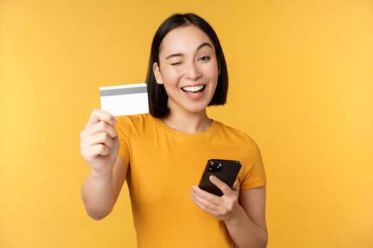 Joyful asian girl smiling, showing credit card and smartphone, recommending mobile phone banking, standing against yellow background.