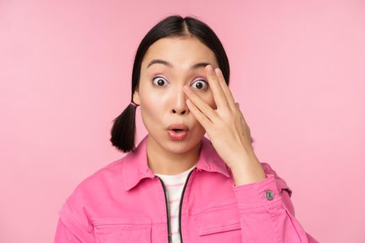 Close up portrait of young asian girl looking surprised, express amazement and wonder, peeking through fingers, standing over pink background.