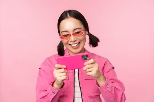 Portrait of happy asian girl playing on smartphone, watching videos on mobile phone app, standing over pink background.
