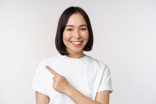 Close up of young japanese woman smiling, pointing finger left at copy space, showing announcement or advertisment banner, standing over white background.