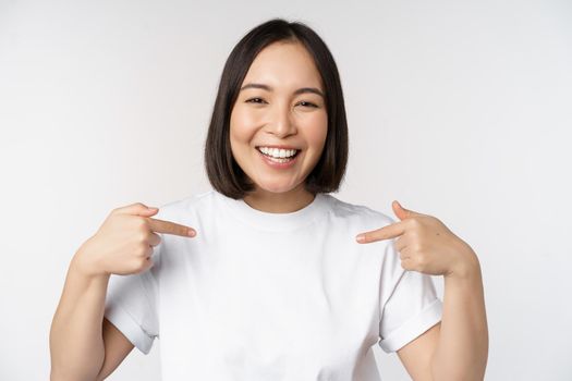 Happy and confident asian woman, student smiling and pointing at herself, self-promoting, showing logo on t-shirt, standing over white background.