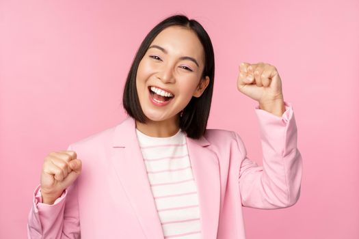 Enthusiastic asian corporate woman, businesswoman raising hand up and cheering, triumphing, winning and celebrating, standing over pink background.