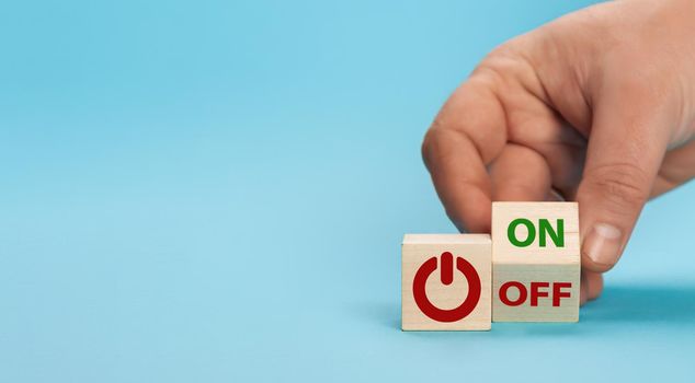 Hand turns a dice and changes the word off to on. Switch button on a wooden cube with inscriptions on and off word on a blue background. Start-stop button