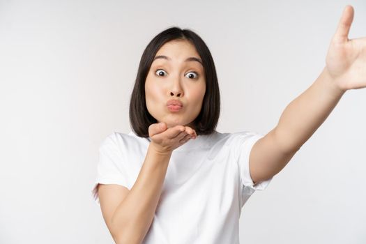 Beautiful young asian woman smiling, looking at camera, holding device, taking selfie, video chat, standing in tshirt over white background.