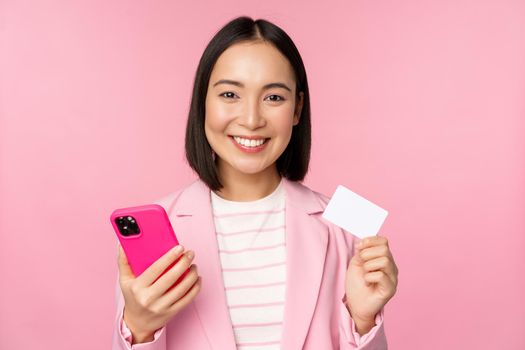 Image of smiling happy asian businesswoman showing credit card, paying online on smartphone application, order with mobile phon, standing against pink background.