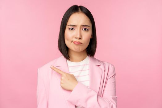 Skeptical businesswoman, pointing finger left, sulking and grimacing upset, showing smth with disappointment, standing over pink background.