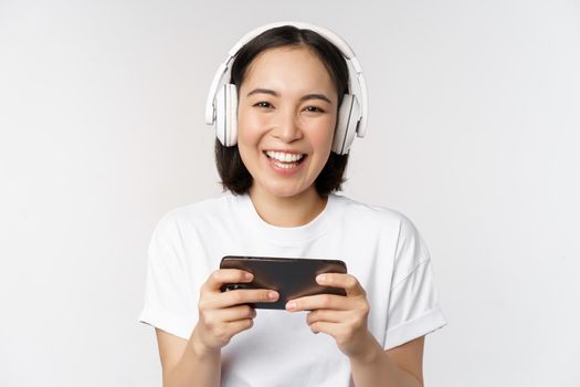 Happy asian girl gamer, playing on mobile phone, watching on smartphone, wearing headphones, standing over white background.