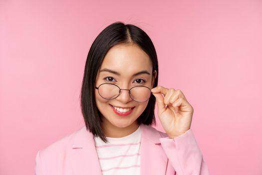 Portrait of asian businesswoman in glasses, looking intrigued at camera and smiling, professional saleswoman staring with interest, pink background.