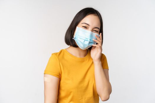 Covid-19, vaccination and healthcare concept. Cute asian girl in medical face mask, showing band aid after coronavirus vaccination, standing over white background.